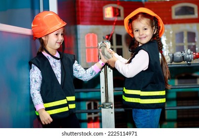 Two little girls dressed as firefighters play role-playing games and water a toy house with a hose, girls in a hard hat, children's games.
