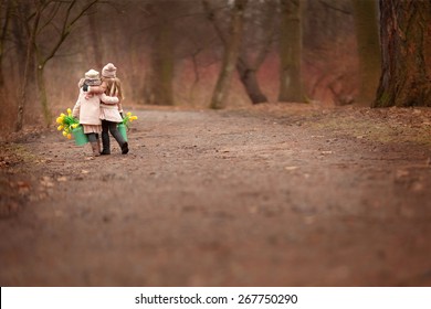 Two Little Girls Friends  Two Green Cans With Yellow Flowers Tulips Are Walking On The Road Hugging In The Park Or Forest 