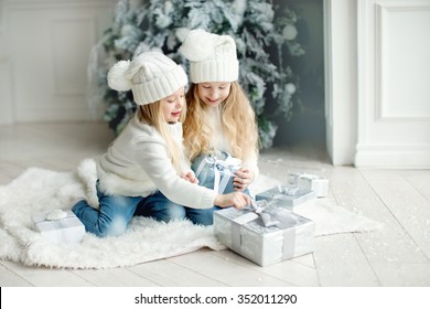two little girls friends or sisters with long blond hair in white hats and white cardigans and jeans playing andand smiling near the white christmas tree with presents at winter time