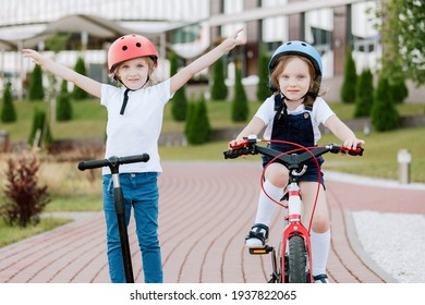 Two little girls having fun on bicycle and scooter. Cheerful sisters in helmets riding outdoors.