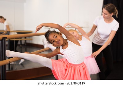 Two little girls practicing choreographic elements on ballet barre with help of teacher in dance school