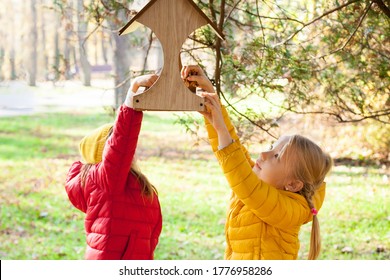 Two Little Girls Putting Food For Birds At Birdhouse  In The Forest On Warm Autumn Day. Children Taking Care Of Animals Outdoor. Outdoor Recreation And Awesome Adventures With Kids In Fall