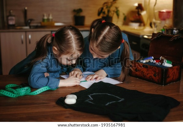Two little girls sew a mask at home. They draw a pattern\
on paper. Nearby is fabric and multi-colored threads. Seamstress\
