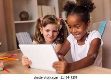 Two little girls sitting at a desk, playing video games on a tablet computer and having fun. Focus on the girl on the left