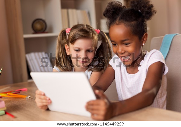 2人の小さな女の子が机に座り、タブレットPCでビデオゲームをして楽しむ。左の女の子に集中する