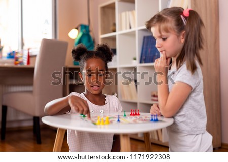 Two little girls sitting in a playroom, playing a ludo board game. Focus on the girl on the left
