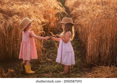 Two Little Girls In Straw Hats And Pink Dresses Are Having Fun In Wheat Field. Girlfriends Tickle Each Other's Nose With Spikelets Of Rye. Sister In Yellow Rubber Boots, The Other Barefoot In The Mud