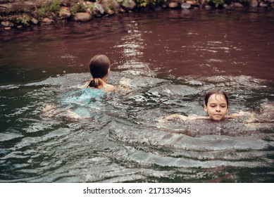 Two little girls in summer dresses swim in a pond outdoors. Children rest in the village, summer vacation. Carefree childhood. The concept of relaxation and fun. Children swim in the pool.