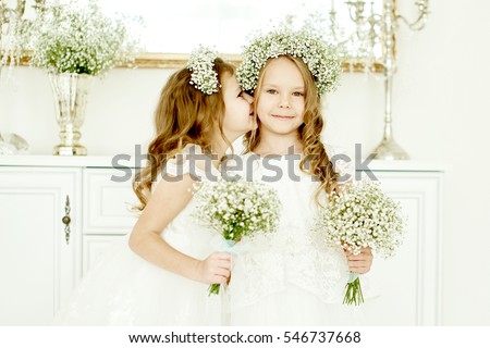 two little girls in wedding dresses standing at the dresser with a big mirror and held bunches of flowers