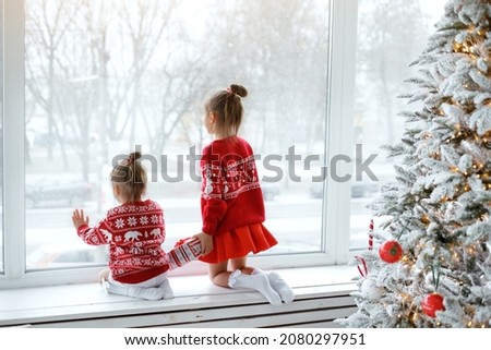 Two little happy girls are watching the first snow through the window, sitting on the windowsill. Kids in Christmas red sweaters next to a decorated Christmas tree are waiting for Christmas Eve. xmas 