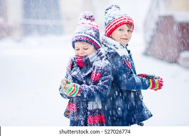 Two Little Kid Boys In Colorful Clothes Playing Outdoors During Snowfall. Active Leisure With Children In Winter On Cold Days. Happy Siblings And Twins Having Fun With Snow