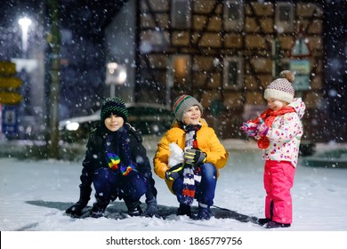 Two Little Kid Boys And Cute Toddler Girl Sitting Together Playing With Snow On Winter Night. Siblings, Brothers And Baby Sister Enjoying Strong Snowfall. Active Fun For Family Vacation