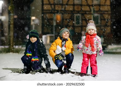 Two Little Kid Boys And Cute Toddler Girl Sitting Together Playing With Snow On Winter Night. Siblings, Brothers And Baby Sister Enjoying Strong Snowfall. Active Fun For Family Vacation