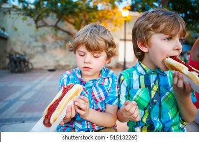 Two little kid boys eating hot dogs outdoors. Siblings enjoying their meal. Hotdog as nhealthy food for children.