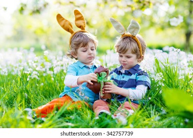 Two Little Kid Boys Playing Together With Chocolate Bunny. Children Making Easter Egg Hunt And Wearing Bunny Ears. Friends Having Fun Outdoors On Sunny Day.