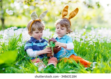 Two Little Kid Boys Playing Together With Chocolate Bunny. Children Making Easter Egg Hunt And Wearing Bunny Ears. Friends Having Fun Outdoors On Sunny Day.