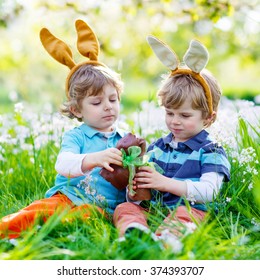 Two Little Kid Boys Playing Together With Chocolate Bunny. Children Making Easter Egg Hunt And Wearing Bunny Ears. Friends Having Fun Outdoors On Sunny Day.