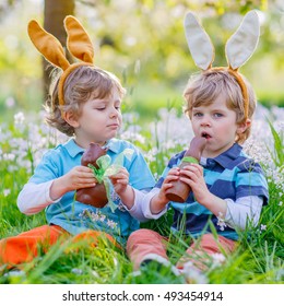 Two Little Kid Boys Playing Together With Chocolate Bunny. Children, Twins Making Easter Egg Hunt And Wearing Bunny Ears. Friends Having Fun Outdoors On Sunny Day.