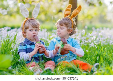 Two Little Kid Boys Playing Together With Chocolate Bunny. Children, Twins Making Easter Egg Hunt And Wearing Bunny Ears. Friends Having Fun Outdoors On Sunny Day.