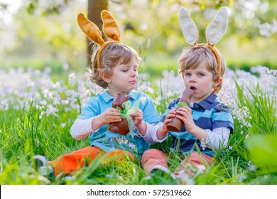 Two Little Kid Boys Playing Together With Chocolate Bunny. Children, Twins Making Easter Egg Hunt And Wearing Bunny Ears. Friends Having Fun Outdoors On Sunny Day.