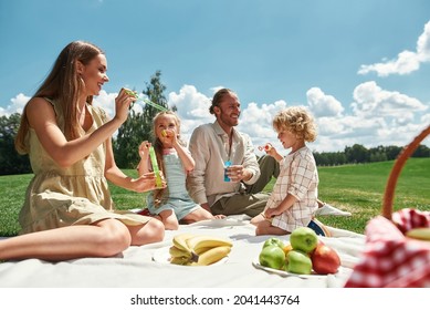 Two Little Kids Blowing Soap Bubbles While Spending Time With Their Parents, Family Having Picnic In Nature On A Summer Day. Leisure, Summer, Childhood Concept