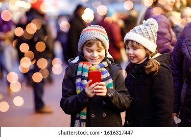 Two Little Kids, Boy And Girl Having Fun On Traditional Christmas Market During Strong Snowfall With Taking Photos With Mobile Smartphone Or Phone. Happy Children, Siblings And Best Friends