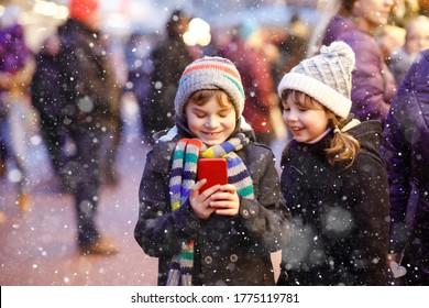 Two Little Kids, Boy And Girl Having Fun On Traditional Christmas Market During Strong Snowfall With Taking Photos With Mobile Smartphone Or Phone. Happy Children, Siblings And Best Friends