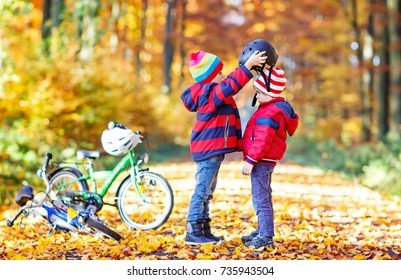 Two Little Kids Boys, Best Friends In Autumn Forest. Older Brother Helping Younger Child Putting Safe Helmet Before Cycling On Sunny Fall Day In Nature. Safety, Sports, Leisure With Kids Concept.