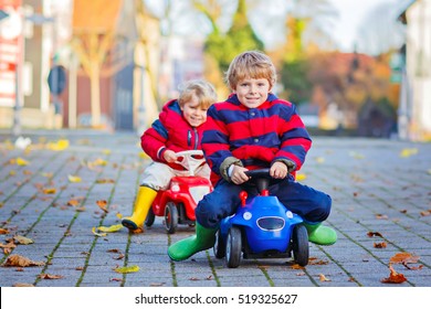 Two Little Kids Boys In Colorful Clothes And Rain Boots Driving Toy Cars. Twins Making Competition, Outdoors. Active Leisure For Children On Autumn Day