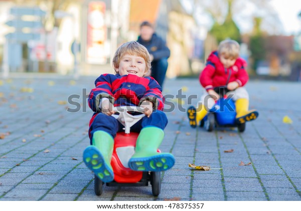Two little kids boys in\
colorful clothes and young dad driving toy cars. Father and sons\
making competition, outdoors. Active leisure for children on autumn\
day