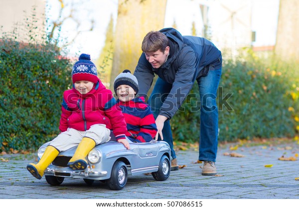 Two little kids boys in colorful\
clothes and young dad driving old car. Father and sons making\
competition, outdoors. Active leisure for children on autumn\
day