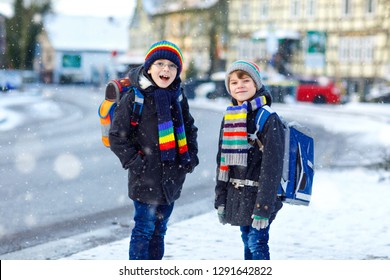 Two Little Kids Boys Of Elementary Class Walking To School During Snowfall. Happy Children Having Fun And Playing With First Snow. Siblings Ans Friends With Backpack In Colorful Winter Clothes.