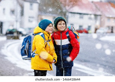 Two Little Kids Boys Of Elementary Class Walking To School During Snowfall. Happy Children Having Fun And Playing With First Snow. Siblings And Best Friends With Backpack In Colorful Winter Clothes.