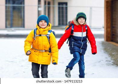 Two Little Kids Boys Of Elementary Class Walking To School During Snowfall. Happy Children Having Fun And Playing With First Snow. Siblings And Best Friends With Backpack In Colorful Winter Clothes.