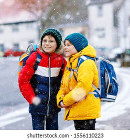 Two Little Kids Boys Of Elementary Class Walking To School During Snowfall. Happy Children Having Fun And Playing With First Snow. Siblings And Best Friends With Backpack In Colorful Winter Clothes.