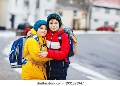 Two Little Kids Boys Of Elementary Class Walking To School During Snowfall. Happy Children Having Fun And Playing With First Snow. Siblings And Best Friends With Backpack In Colorful Winter Clothes.