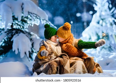 Two little kids celebrating Christmas outdoors holding sparklers in hands. Children wrapped themselves in warm woolen plaid. Fairy winter night, snow-covered Christmas trees around. - Powered by Shutterstock