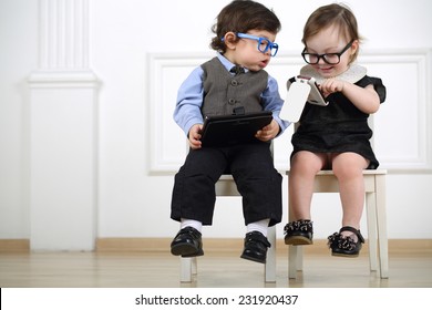 Two Little Kids In Glasses Sitting On White Chairs: Boy With Tablet Computer, Girl With Mobile Phone