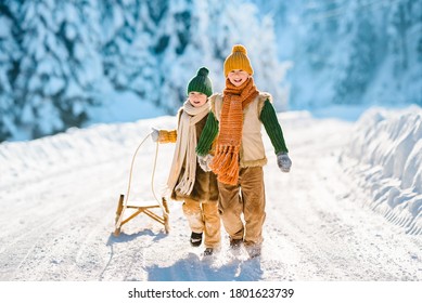 Two Little Kids Have Fun In The Beautiful Winter Nature With Snow-covered Trees. Children Walk Along A Snowy Road Pulling A Sled. Winter Knitted Wool Retro Clothes.