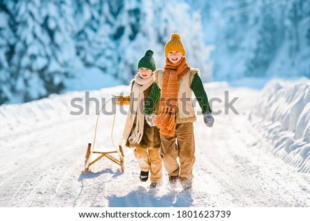 Two little kids have fun in the beautiful winter nature with snow-covered trees. Children walk along a snowy road pulling a sled. Winter knitted wool retro clothes.