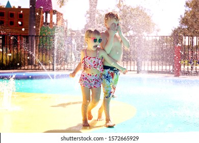 Two Little Kids Are Running And Playing Outside In The Water Fountains At A Community Splash Park On A Sumer Day.