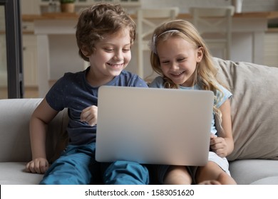 Two little kids siblings seated on sofa in living room holding on lap laptop play online game, watch cartoons spend weekend using electronic device, bad habit, generation addicted with gadgets concept - Powered by Shutterstock