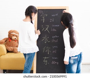 Two Little Kids Standing Looking At Blackboard Writing Chinese Text. Asian Small Girls Study Together At Home To Learn Chinese Language And Write On Chalkboard. New Normal Homeschool Learning Concept.