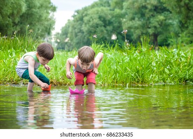 Two Little Sibling Brothers Playing With Paper Boats In A River Or Lake On Warm And Sunny Summer Day. Active Leisure For Children. Kid Boy Best Friends Having Fun Together Outdoors.