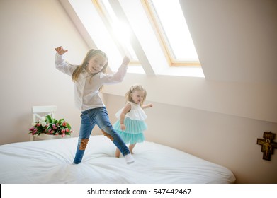 Two little sisters dance on a big bed. To girls it is very cheerful. On a white chair the big beautiful bouquet of tulips lies. From a window the sunlight flows.