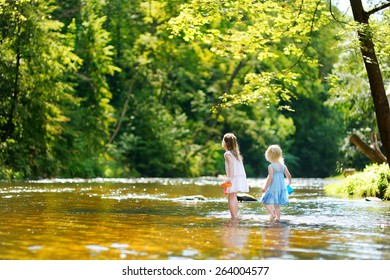 Two little sisters playing with paper boats by a river on warm and sunny summer day
