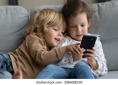 Two little toddler preschooler sibling gen Z kids using online parental control app on mobile phone, playing video game for children, sharing digital gadget, relaxing on comfortable couch at home