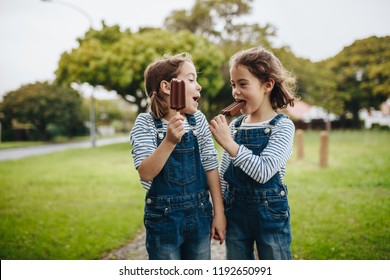 Two little twin sisters in identical clothes standing outdoors and eating chocolate ice cream candy. Two little girls enjoying eating candy icecream outdoors.