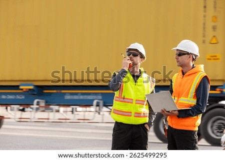 Two logistics workers in high-visibility vests and hard hats stand near a large shipping container, one using a walkie-talkie and the other holding a laptop, coordinating freight operations at a port.