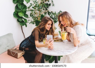 Two long-haired girls resting in cafe with modern interior and laughing. Indoor portrait of funny smiling ladies in trendy clothes making selfie while drink fruit cocktails.  - Powered by Shutterstock